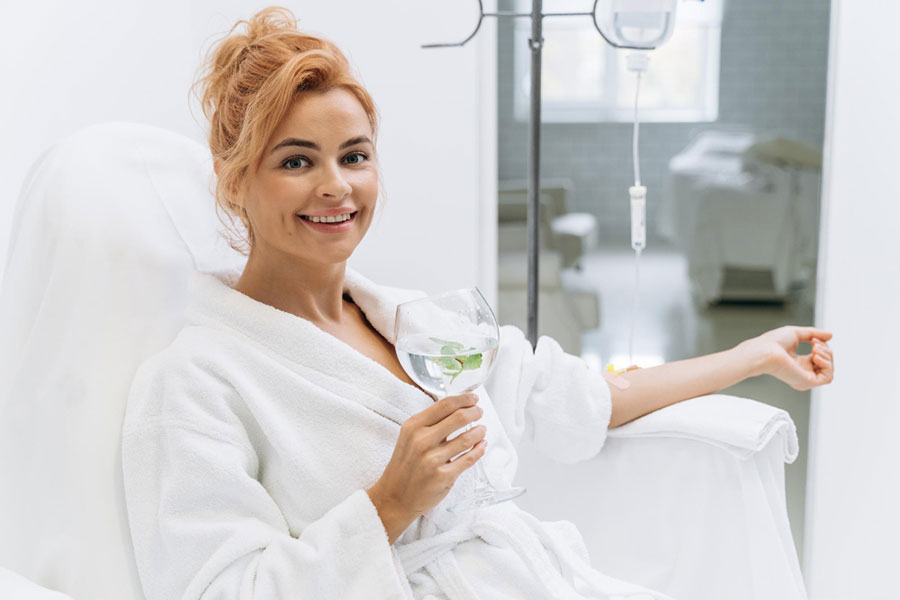 happy-woman-in-white-bathrobe-holding-glass-of-lemon-beverage-and-smiling-while-sitting-in-armchair-and-receiving-iv-infusion iv therapy miami